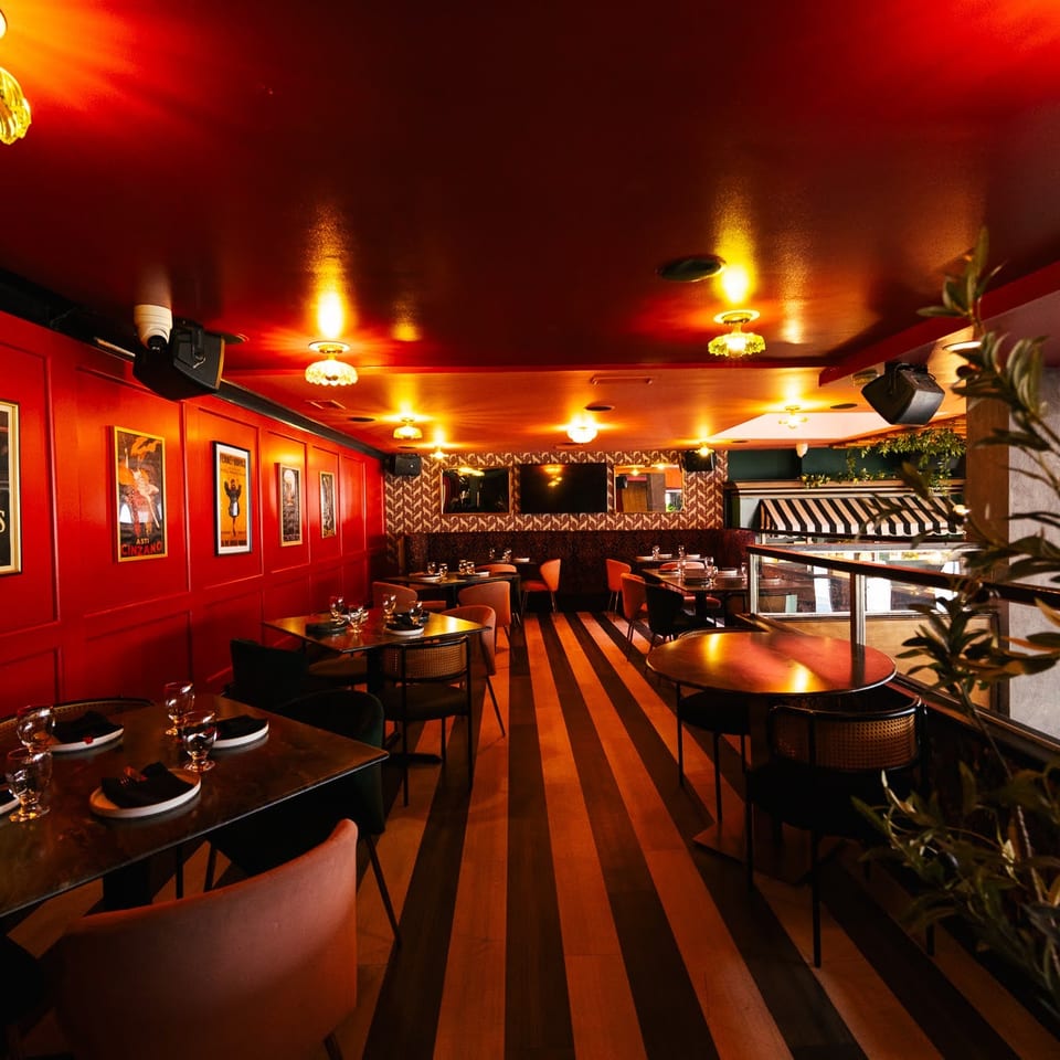 Warmly lit restaurant interior featuring red walls, striped floor, set tables, and vintage posters.