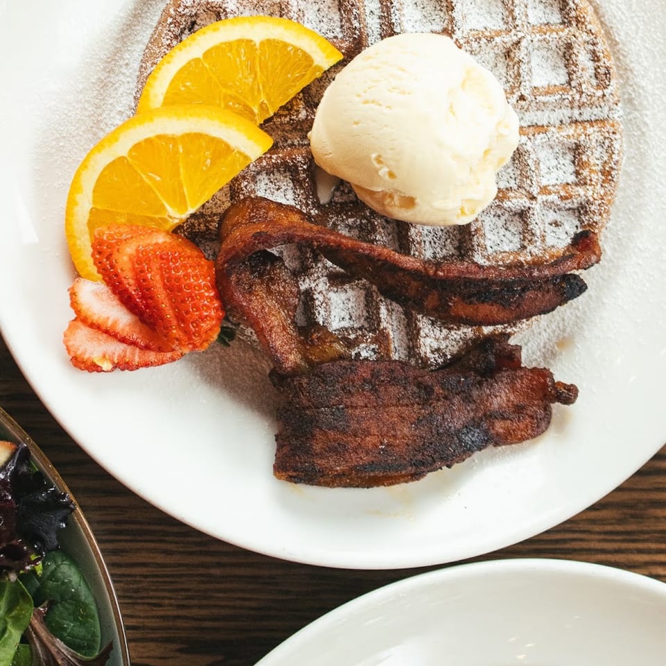 Powdered sugar waffle with ice cream, orange slices, strawberries, and crispy bacon on a white plate.