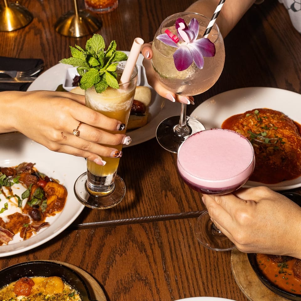 Four people clinking colorful cocktails and drinks over a table laden with various dishes.