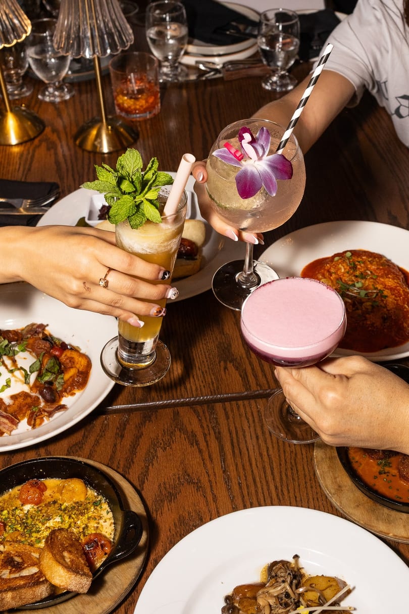 Four people clinking colorful cocktails and drinks over a table laden with various dishes.