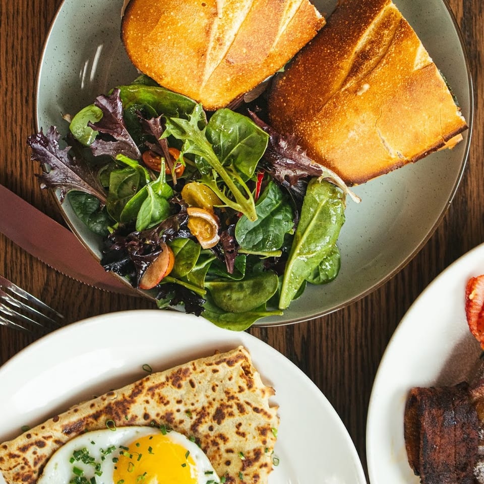 Overhead view of a sandwich with salad, a crepe with fried egg, and bacon on a wooden table.
