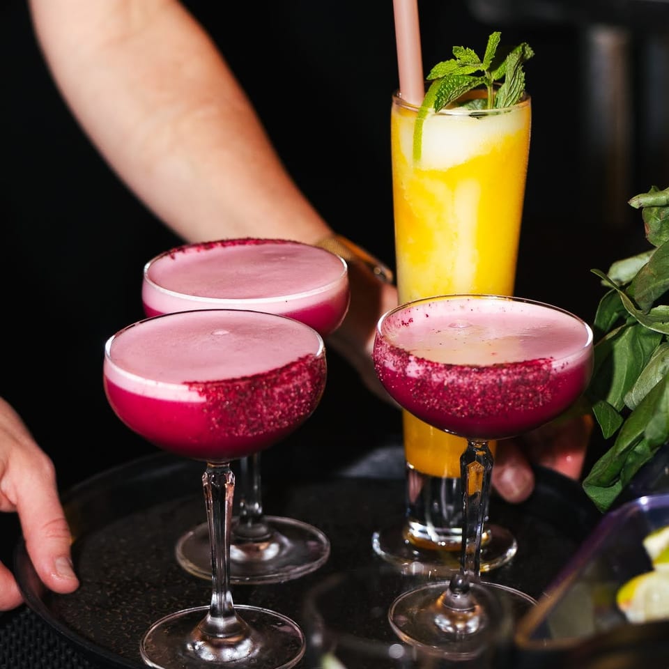A server's hands hold a tray with three cocktails: two pink, one yellow with mint.