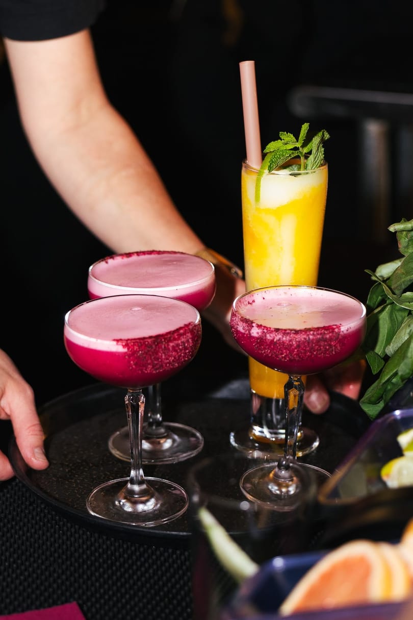 A server's hands hold a tray with three cocktails: two pink, one yellow with mint.