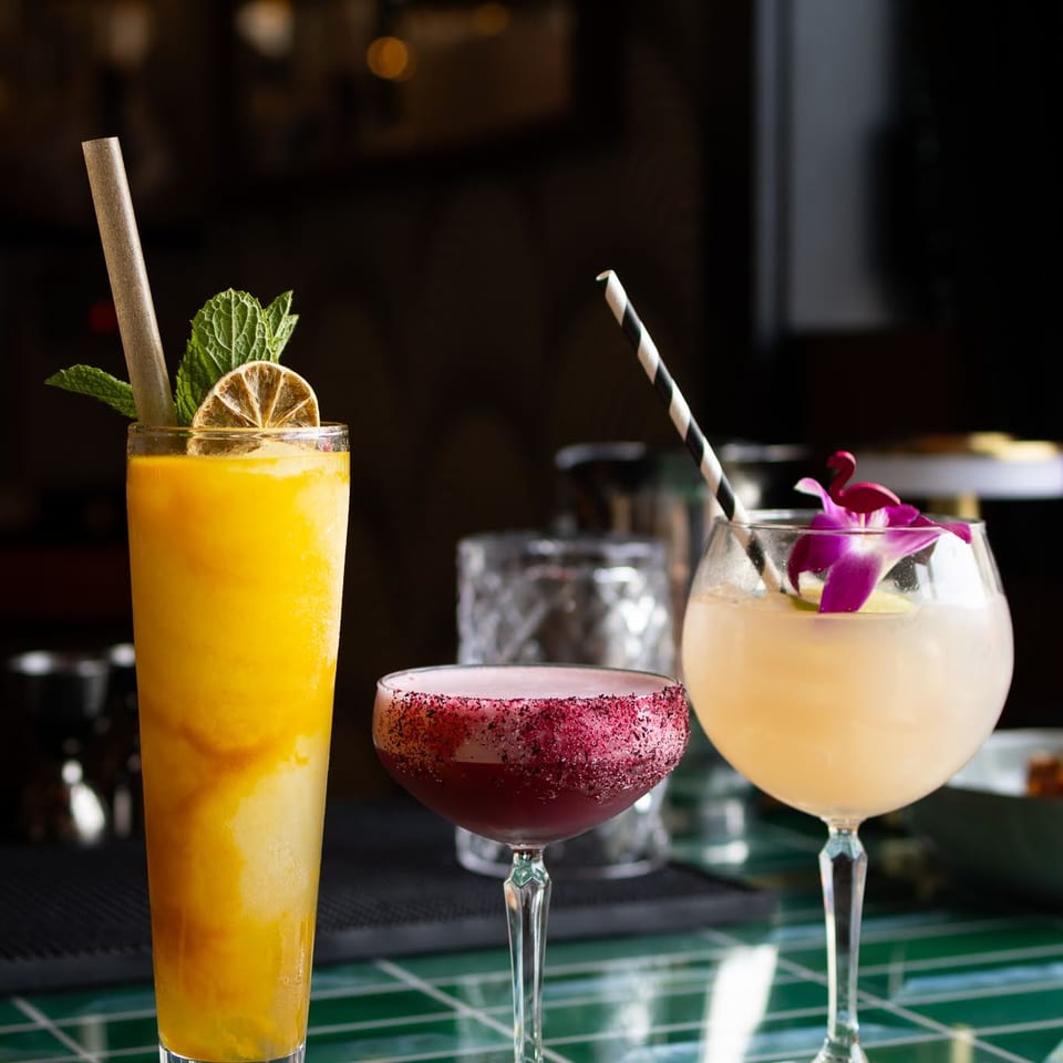 Three colorful cocktails, including a mango smoothie, on a green tiled bar top.