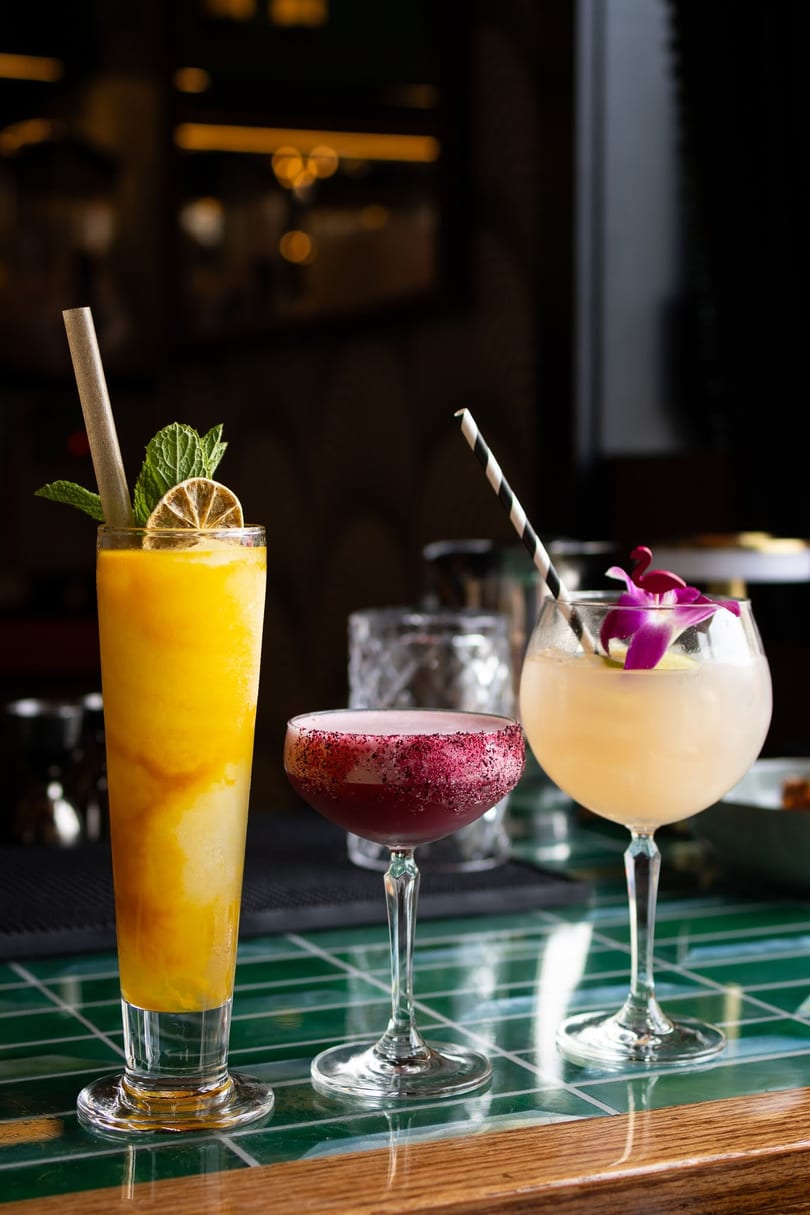 Three colorful cocktails, including a mango smoothie, on a green tiled bar top.