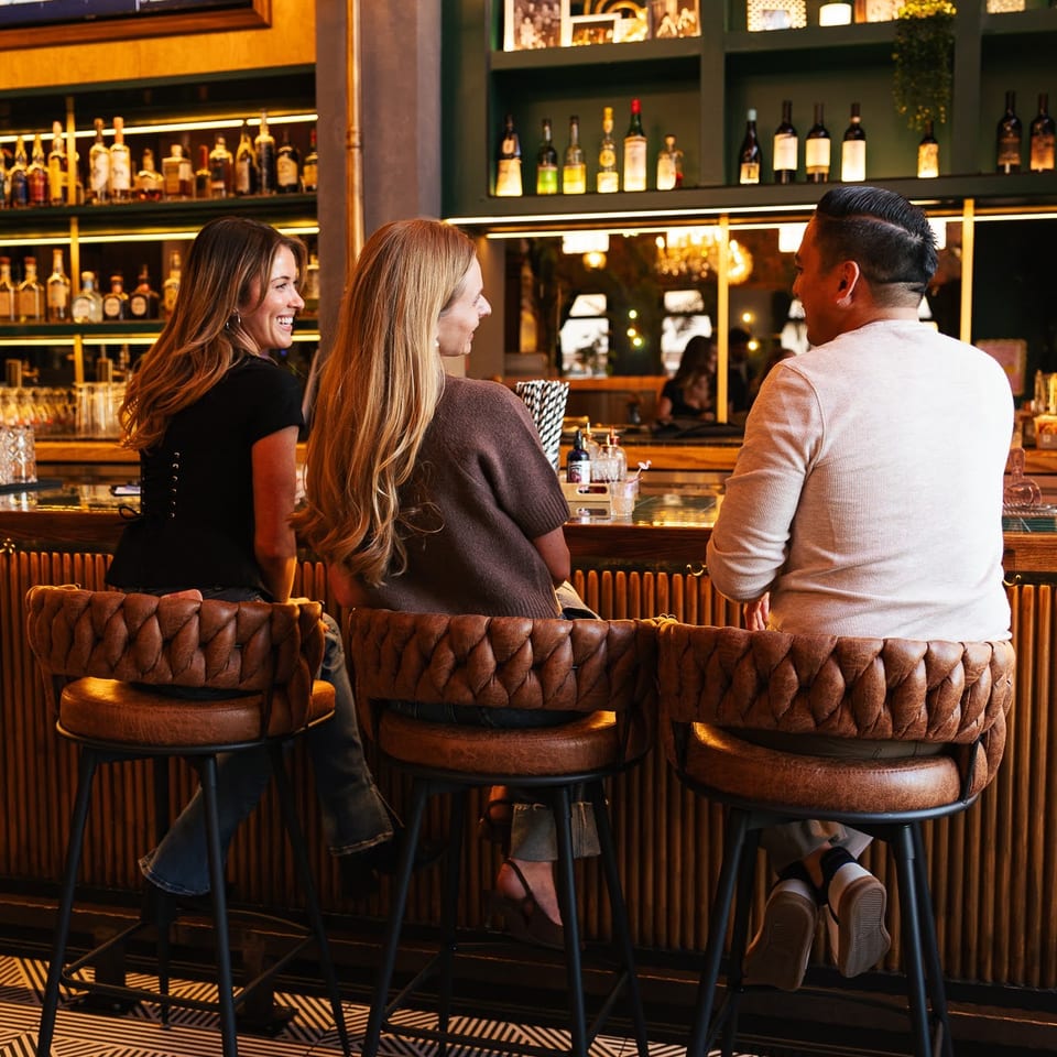 Three people sit at a stylish bar with a geometric floor and well-stocked shelves.