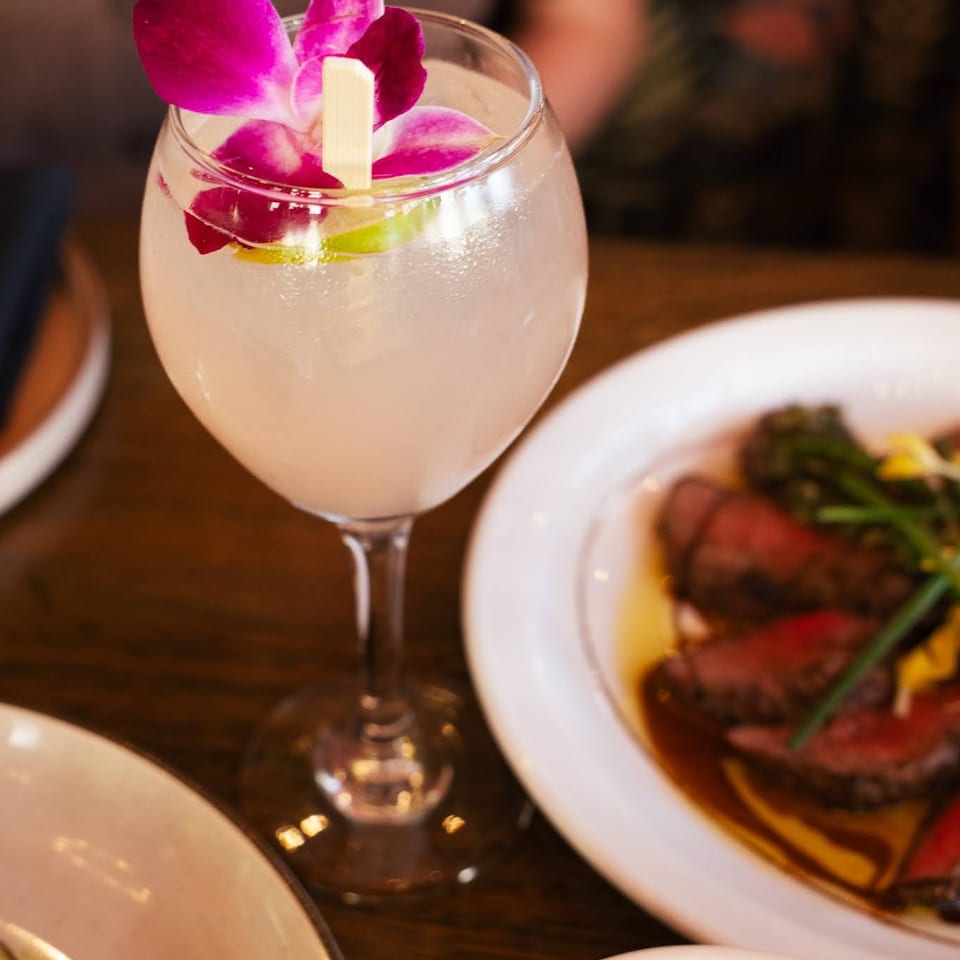 A cocktail garnished with a purple orchid and lime, next to plates of sliced steak and appetizers.