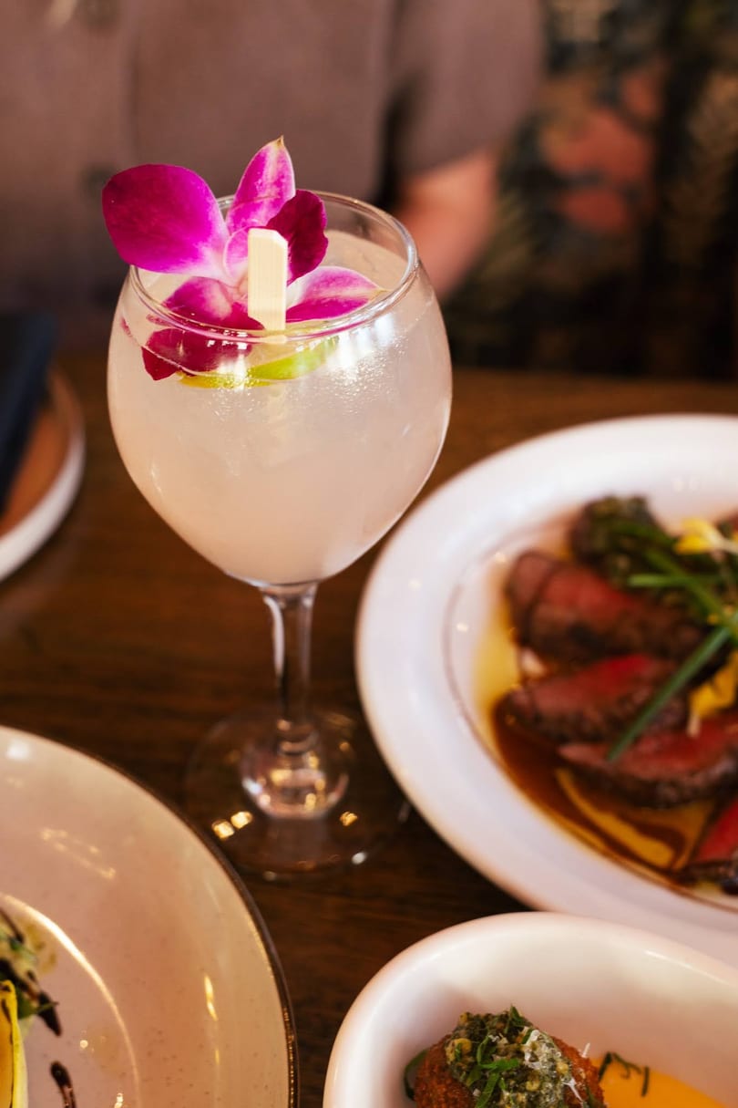 A cocktail garnished with a purple orchid and lime, next to plates of sliced steak and appetizers.