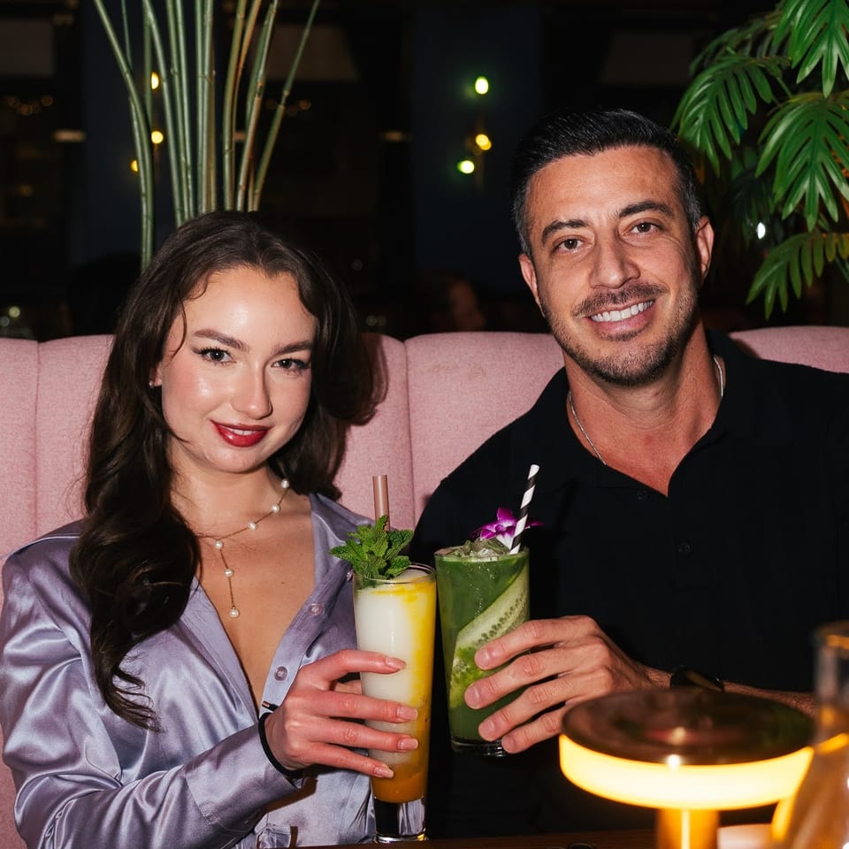 A smiling couple holding colorful cocktails in a dimly lit restaurant with tropical plants.