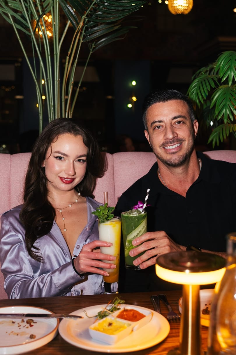 A smiling couple holding colorful cocktails in a dimly lit restaurant with tropical plants.