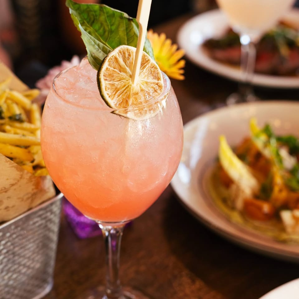 Pink cocktail with dried lime and basil garnish, surrounded by food on a restaurant table.