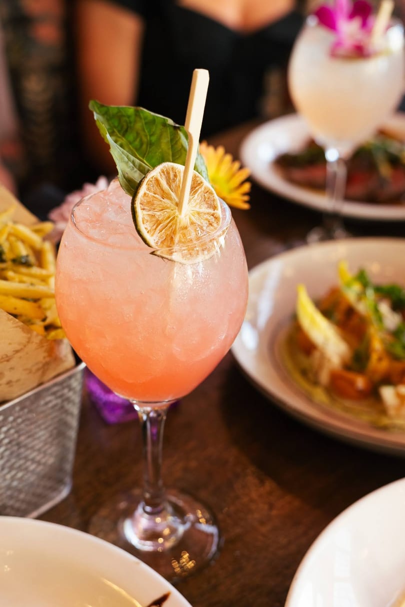 Pink cocktail with dried lime and basil garnish, surrounded by food on a restaurant table.